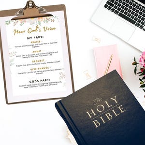 以下が含まれることがあります： 「Hear God's Voice」のテキストと指示のリストが書かれたクリップボード、金色の文字が入った黒い聖書、ラップトップ、ピンクのノート、金のペン、白い表面にピンクの花。