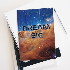 Op de afbeelding: Een spiraalgebonden notitieboek met een galaxy-thema op de omslag, met een nevel met sterren en de tekst "DREAM BIG" in het zwart.