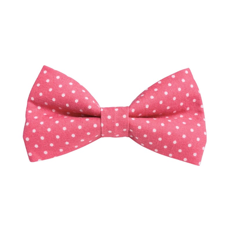 May include: A pink bow tie with a white polka dot pattern. The bow tie is made of fabric and is tied in a classic bow shape. The bow tie is a solid pink color with evenly spaced white polka dots.