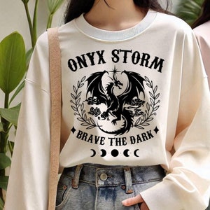 May include: White long-sleeved sweatshirt with a black graphic of a dragon with the text "ONYX STORM" and "BRAVE THE DARK" and three black crescent moons.