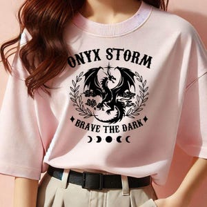 May include: Pink t-shirt with a black graphic of a dragon with the text "ONYX STORM" and "BRAVE THE DARK" with a crescent moon design.