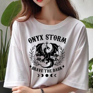 May include: White t-shirt with a black graphic of a dragon with the text "ONYX STORM" above and "BRAVE THE DARK" below. The graphic also includes a star and a crescent moon.