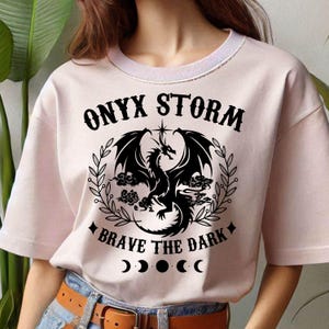 May include: A pink t-shirt with a black dragon graphic and the text "ONYX STORM" and "BRAVE THE DARK". The shirt also has a crescent moon graphic.