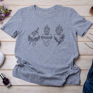 Puede incluir: Camiseta gris jaspeado con tres dise&ntilde;os de coraz&oacute;n negros. Cada coraz&oacute;n est&aacute; adornado con un s&iacute;mbolo diferente: una espada, una corona de espinas y un motivo floral. La camiseta est&aacute; hecha de tela suave.