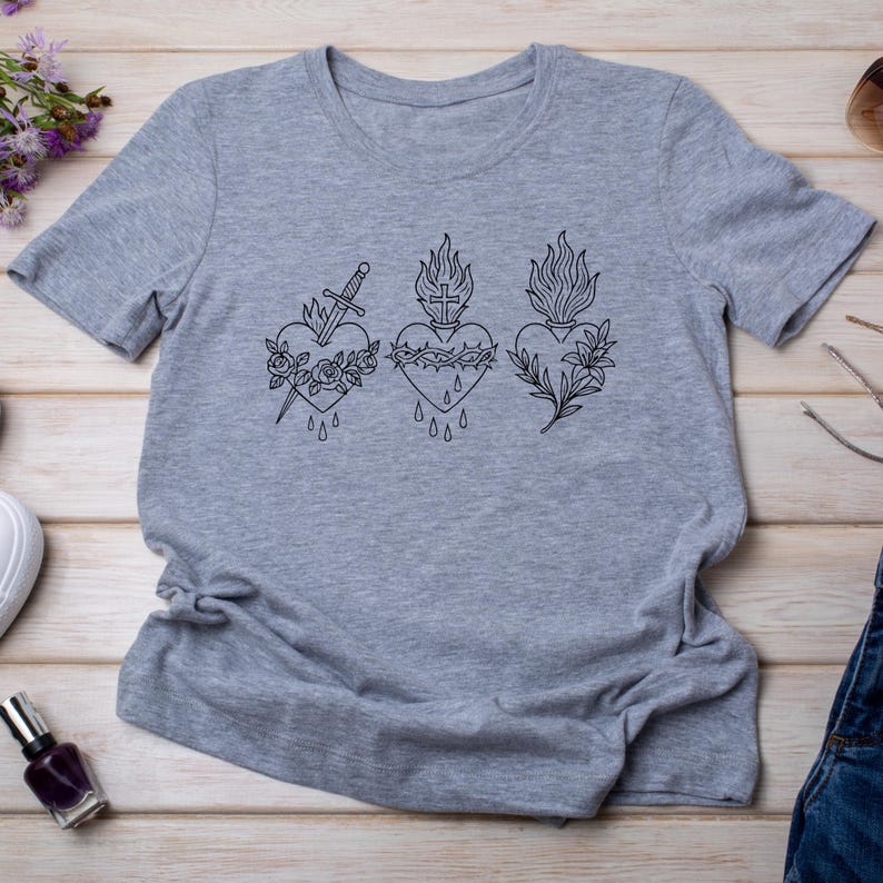 Puede incluir: Camiseta gris jaspeado con tres dise&ntilde;os de coraz&oacute;n negros. Cada coraz&oacute;n est&aacute; adornado con un s&iacute;mbolo diferente: una espada, una corona de espinas y un motivo floral. La camiseta est&aacute; hecha de tela suave.