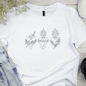 Puede incluir: Camiseta blanca con tres dise&ntilde;os de coraz&oacute;n negros. Cada coraz&oacute;n presenta un dise&ntilde;o diferente: una espada y rosas, una corona de espinas y l&aacute;grimas, y una ramita floral. La camiseta est&aacute; hecha de un material suave y c&oacute;modo.