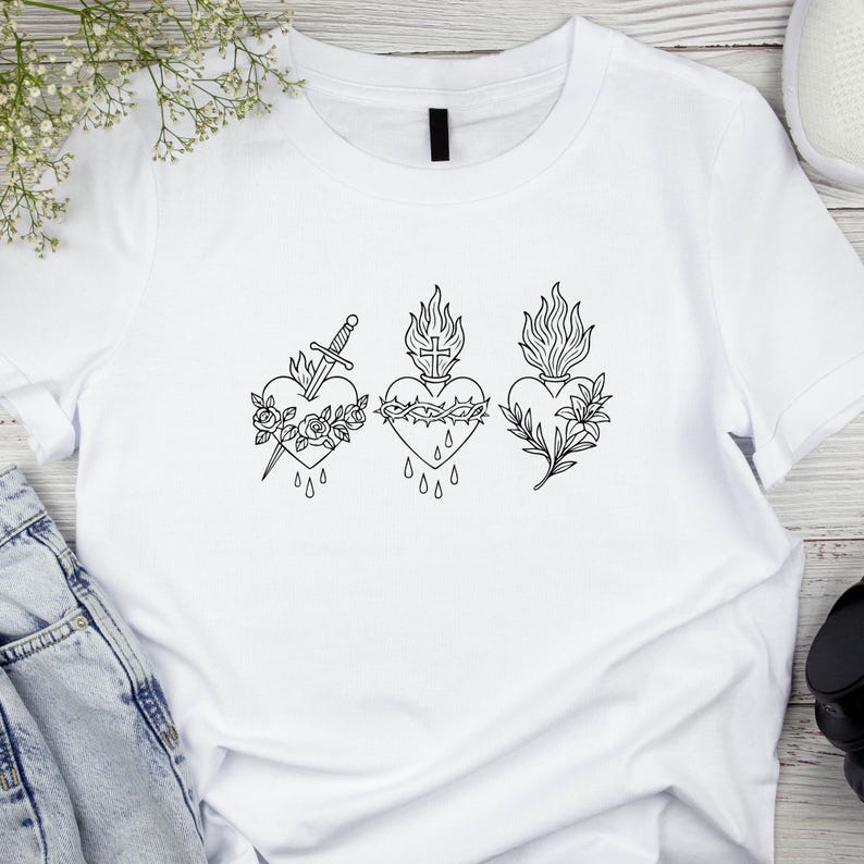 Puede incluir: Camiseta blanca con tres dise&ntilde;os de coraz&oacute;n negros. Cada coraz&oacute;n presenta un dise&ntilde;o diferente: una espada y rosas, una corona de espinas y l&aacute;grimas, y una ramita floral. La camiseta est&aacute; hecha de un material suave y c&oacute;modo.