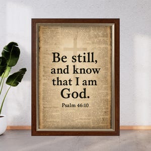 May include: Framed print with the text "Be still, and know that I am God." in black, with a cross graphic, set against a vintage newspaper background. The frame is a dark brown color.