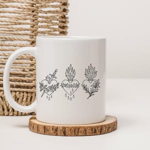Puede incluir: Taza de cer&aacute;mica blanca con tres corazones negros dibujados a l&iacute;nea. El primer coraz&oacute;n tiene una espada y rosas, el segundo una corona de espinas y una cruz, y el tercero llamas y hojas. La taza est&aacute; sobre un posavasos de madera.