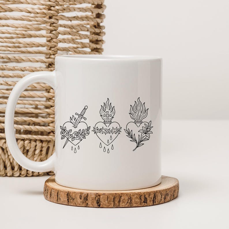 Puede incluir: Taza de cer&aacute;mica blanca con tres corazones negros dibujados a l&iacute;nea. El primer coraz&oacute;n tiene una espada y rosas, el segundo una corona de espinas y una cruz, y el tercero llamas y hojas. La taza est&aacute; sobre un posavasos de madera.