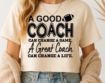 May include: Cream-colored crop top with the text "A GOOD COACH CAN CHANGE A GAME, A Great Coach CAN CHANGE A LIFE." A black football graphic is above the word "COACH". The shirt is paired with blue jeans.