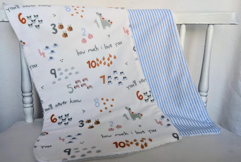 Pode incluir: Um cobertor de beb&ecirc; branco com n&uacute;meros coloridos, insetos e uma estampa de dinossauro, junto com o texto "how much I love you" e "you'll never know". O verso apresenta listras azuis e brancas.