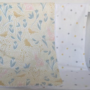 Pode incluir: Duas mantas de beb&ecirc; sobre uma cadeira branca. Uma manta apresenta um fundo creme com um padr&atilde;o de p&aacute;ssaros, coelhos e flores em azul, rosa e dourado. A outra manta &eacute; branca com bolinhas cinza e amarelas.