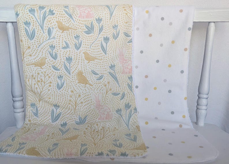 Pode incluir: Duas mantas de beb&ecirc; sobre uma cadeira branca. Uma manta apresenta um fundo creme com um padr&atilde;o de p&aacute;ssaros, coelhos e flores em azul, rosa e dourado. A outra manta &eacute; branca com bolinhas cinza e amarelas.
