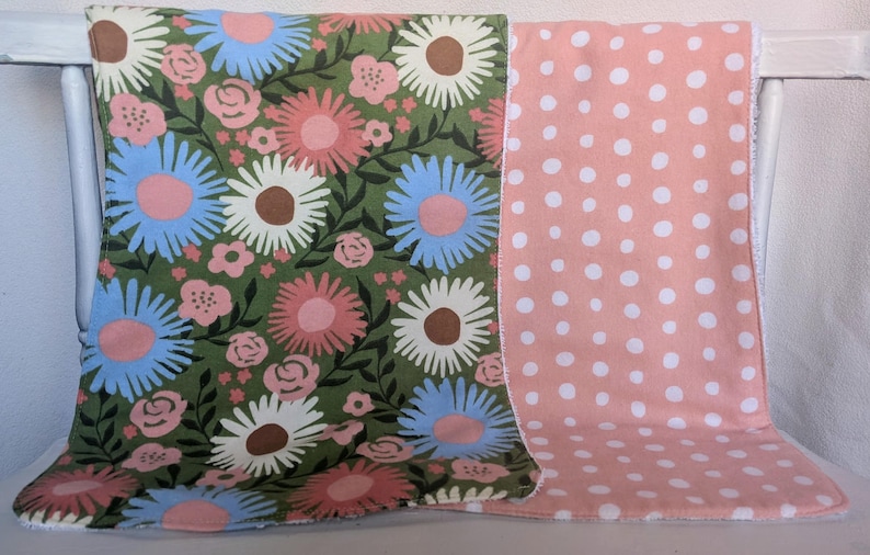 Pode incluir: Um cobertor revers&iacute;vel para beb&eacute;. Um lado tem um design floral com margaridas rosa, azuis e brancas sobre um fundo verde. O outro lado &eacute; rosa com bolinhas brancas. O cobertor &eacute; projetado para beb&eacute;s.