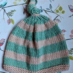 May include: A green and pink striped knitted hat with a knotted top. The hat has a pink brim.
