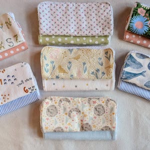 Pode incluir: Uma cole&ccedil;&atilde;o de sete itens de tecido retangulares, cada um com um design de padr&atilde;o &uacute;nico. Os padr&otilde;es incluem estampas florais, de baleias e alfab&eacute;ticas, com v&aacute;rias combina&ccedil;&otilde;es de cores como azul, rosa e verde. Os itens parecem estar dobrados e empilhados.