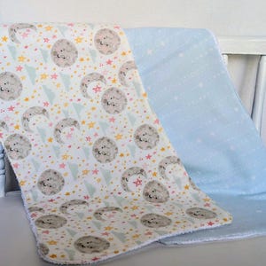 Pode incluir: Duas mantas de beb&eacute;. Uma &eacute; branca com um padr&atilde;o de luas cinzentas, estrelas e tri&acirc;ngulos em amarelo, rosa e azul. A outra &eacute; azul claro liso com estrelas brancas. As mantas est&atilde;o dobradas sobre uma cadeira branca.
