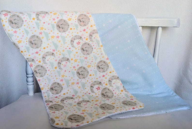 Pode incluir: Duas mantas de beb&eacute;. Uma &eacute; branca com um padr&atilde;o de luas cinzentas, estrelas e tri&acirc;ngulos em amarelo, rosa e azul. A outra &eacute; azul claro liso com estrelas brancas. As mantas est&atilde;o dobradas sobre uma cadeira branca.
