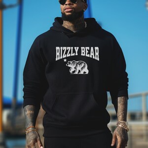 Puede incluir: Sudadera con capucha negra con un gráfico blanco de un oso y el texto "Rizzly Bear".