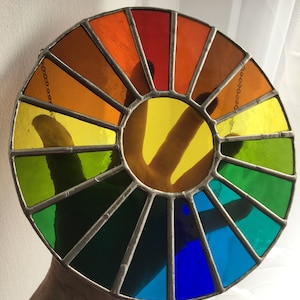 May include: A circular stained glass piece depicting a colour wheel. The design features segments of red, orange, yellow, green, and blue, separated by metal. A hand is visible behind the yellow centre.