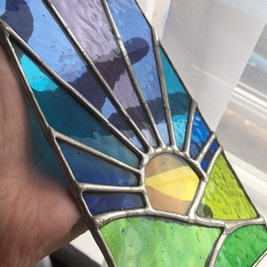May include: A stained glass suncatcher with a sunburst design. It features a palette of blues, purples, yellows, and greens, framed in silver-coloured metal. A decorative piece for windows or walls, reminiscent of a sunrise.