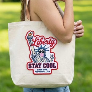 Pode incluir: Uma sacola de lona bege com um gráfico da Estátua da Liberdade usando óculos de sol e soprando uma bolha rosa. O design inclui o texto Liberty, STAY COOL, Since 1776 e Anywhere, USA, com uma borda vermelha ao redor da ilustração.