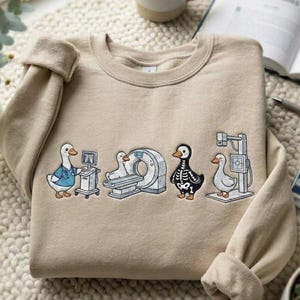Op de afbeelding: Beige crewneck sweatshirt met geborduurde cartoon eenden in medische settings. Eén eend draagt een blauwe jas, een andere heeft een skeletontwerp en anderen staan in de buurt van medische apparatuur. Een mok, boek en plant staan op de achtergrond.