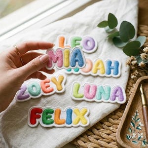 May include: A collection of colorful, puffy, embroidered name patches featuring the names Leo, Mia, Noah, Zoey, Luna, and Felix. Each letter is stitched in a vibrant, solid color with a white border, designed to be applied to fabric items.