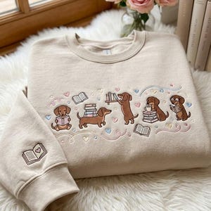 Könnte beinhalten: Beiges Sweatshirt mit gestickten Dackeln, die Bücher lesen. Das Design zeigt mehrere braune Dackel, die mit Büchern, Herzen und pastellfarbenen Akzenten interagieren. Der linke Ärmel hat ein kleines gesticktes Buch mit einem Herzen.