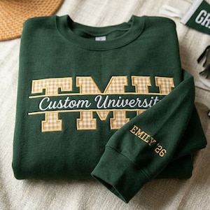 Op de afbeelding: Een donkergroene sweatshirt met aangepaste geborduurde tekst. Het ontwerp bevat de woorden "Custom University" en de initialen "TM" in een bruin geruit patroon. De mouw is geborduurd met "EMILY '26".
