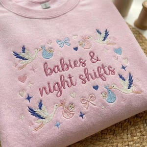 May include: A light pink sweatshirt with embroidered text that reads "babies & night shifts". The design includes storks carrying babies, hearts, bows, and stars in shades of blue, pink, and gold.