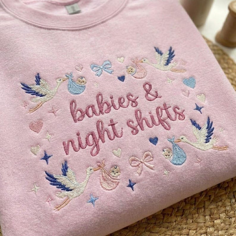 May include: A light pink sweatshirt with embroidered text that reads "babies & night shifts". The design includes storks carrying babies, hearts, bows, and stars in shades of blue, pink, and gold.