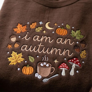 May include: A dark brown sweatshirt featuring embroidered text that reads i am an autumn. The design includes colorful autumn-themed motifs such as orange pumpkins, red and white mushrooms, maple leaves, acorns, a crescent moon, and a mug of cocoa.