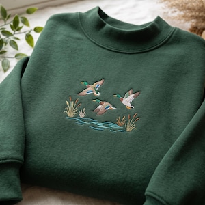 Peut inclure: Un sweat-shirt vert forêt foncé doté d'un col montant et de manches longues. Le centre de la poitrine présente une scène brodée de trois canards colverts en vol au-dessus d'une surface d'eau bleue avec des massettes brunes et jaunes.