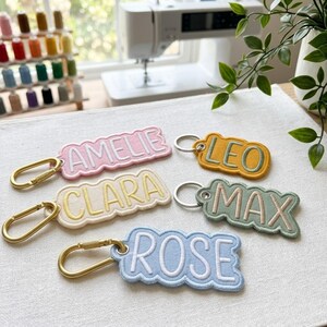 Op de afbeelding: Vijf stoffen sleutelhangers met geschulpte randen en geborduurde namen. Het roze label leest AMELIE, geel leest CLARA, mosterdgeel leest LEO, saliegroen leest MAX en lichtblauw leest ROSE. Elke sleutelhanger heeft een goudkleurige metalen karabijnhaak.