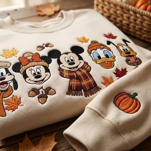 May include: A cream-colored crewneck sweatshirt featuring embroidered character faces including Mickey Mouse, Minnie Mouse, Donald Duck, Pluto, and Goofy. The design includes autumn-themed elements like orange and red maple leaves, acorns, and a pumpkin on the sleeve.