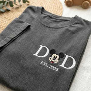 May include: Dark gray t-shirt with white embroidered text and a Mickey Mouse design. The text reads "DAD" with "EST. 2026" below the design. The shirt is folded, with the sleeve visible.