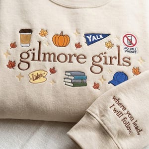 Peut inclure: Un sweat-shirt beige avec une broderie marron indiquant gilmore girls. Le motif inclut une tasse de café, une citrouille, un fanion Yale, un panneau Luke's, des livres, une casquette bleue et un signe no cell phones. La manche porte le texte where you lead, I will follow.