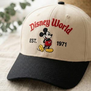 Op de afbeelding: Beige baseballpet met een zwarte klep. De pet heeft een rode borduursel met de tekst "Disney World", een Mickey Mouse-afbeelding en de tekst "EST. 1971". Er zit een zwarte knop bovenop.