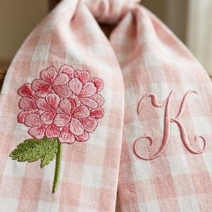 Può includere: Due asciugamani a quadretti rosa e bianchi. Un asciugamano presenta un ricamo di fiore di ortensia rosa con foglie e stelo verdi. L'altro asciugamano ha una lettera "K" ricamata in rosa.