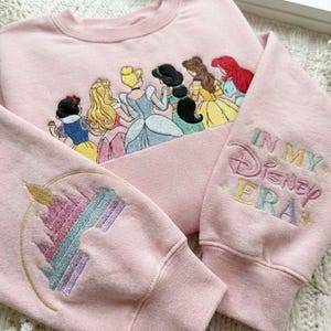 May include: Pink sweatshirt with embroidered Disney princesses across the chest. The sleeve features a castle design and the words "IN MY Disney ERA" in pastel colors. A cozy and colorful garment.