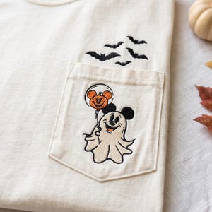 May include: An off-white t-shirt featuring an embroidered pocket design. The pocket displays a ghost character wearing Mickey Mouse ears and holding an orange pumpkin balloon. Above the pocket, four small black bats are embroidered in flight.