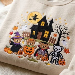 May include: A beige sweatshirt featuring an embroidered Halloween scene with four teddy bears dressed as a witch, a pumpkin, a vampire, and a skeleton. The design includes a black haunted house, a yellow moon with a witch silhouette, and ghosts.
