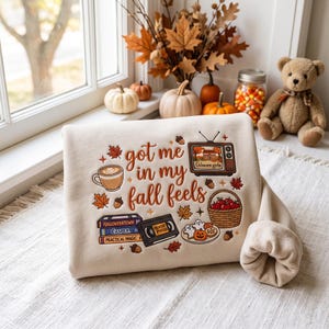 May include: A beige sweatshirt featuring the text "got me in my fall feels" surrounded by illustrations of a coffee cup, a vintage television showing "Gilmore girls", a basket of apples, a stack of VHS tapes, and a plate of cookies.