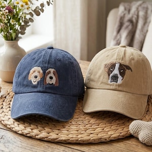 Pode incluir: Dois bonés de beisebol com rostos de cães bordados. Um é azul marinho com dois rostos de cães, e o outro é bege com um rosto de cão. Ambos os bonés têm uma aba curva e um botão na parte superior.