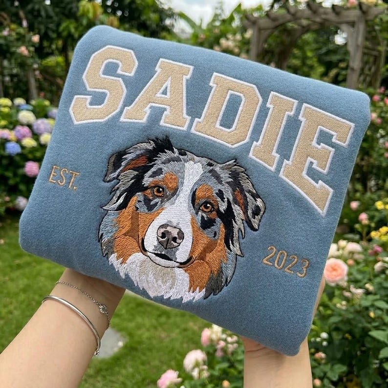 May include: A blue sweatshirt with the name "SADIE" embroidered in tan block letters. Below the name is an embroidered dog portrait with brown, gray, and white fur. The text "EST. 2023" is also embroidered on the sweatshirt.