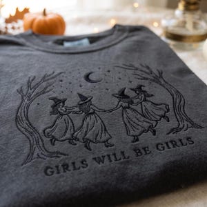 Peut inclure: Un sweat-shirt gris anthracite avec une broderie noire repr&eacute;sentant quatre silhouettes portant des chapeaux de sorci&egrave;re dansant en cercle entre deux arbres sous un croissant de lune et des &eacute;toiles, avec le texte GIRLS WILL BE GIRLS brod&eacute;.