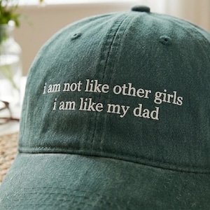 May include: A dark green baseball cap with white embroidered text that reads "I am not like other girls I am like my dad". The cap is made of a soft fabric and has a curved brim.