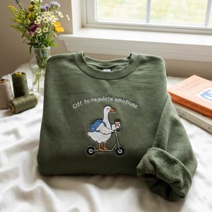 Puede incluir: Una sudadera verde oliva con la frase bordada "Off to regulate emotions". El dise&ntilde;o presenta un ganso montando un scooter con una mochila y una taza de caf&eacute;. La sudadera est&aacute; doblada sobre una superficie blanca.